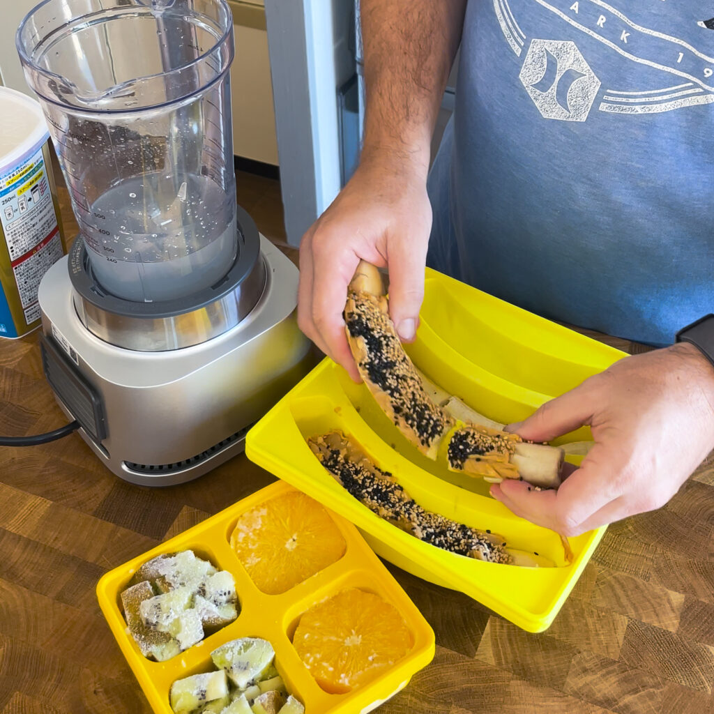Smootheeze banana silicone freezer tray filled with bananas that are topped with peanut butter. A person is putting the bananas in a blender to make a smoothie.
