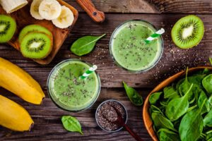 Green smoothies in a glass surrounded by bananas, spinach, and kiwis.