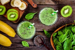 Image of green smoothies in a glass surrounded by bananas, spinach, and kiwis.