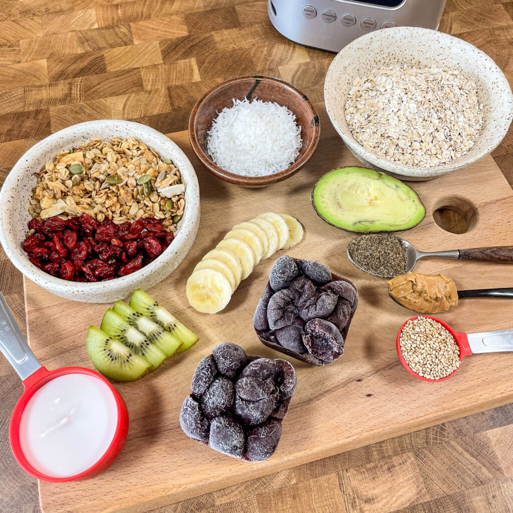 A collection of smoothie bowl ingredients ready to blend, including frozen cherries, bananas, avocado, kiwi, granola, and seeds.