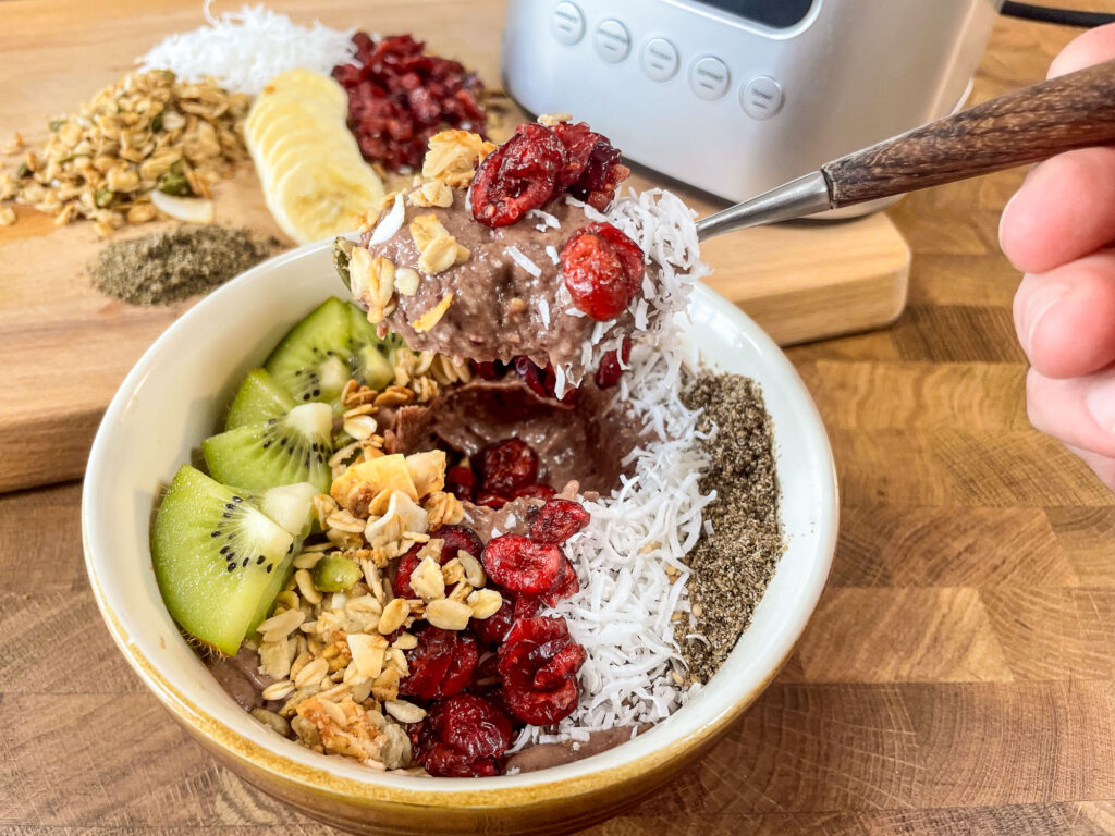 A smoothie bowl made from frozen fruit topped with coconut, granola, kiwi, and chia seeds, positioned on the counter.